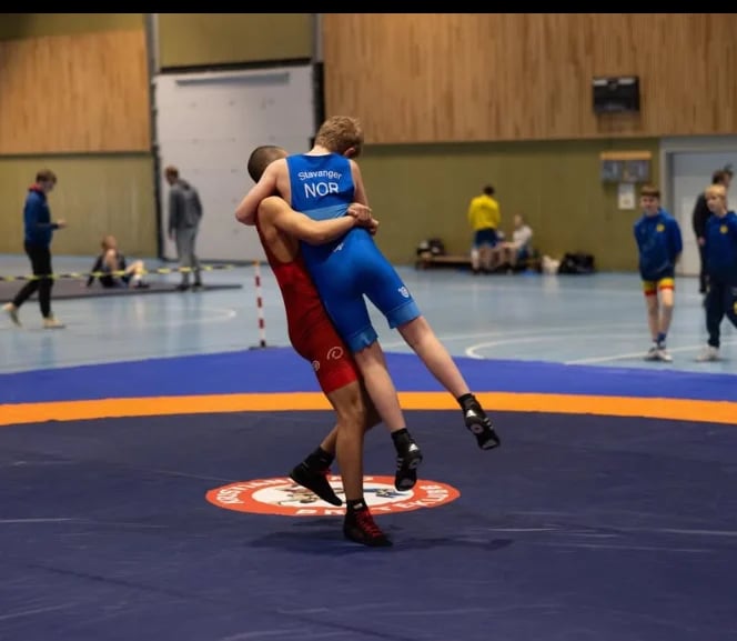 Two wrestlers grappling on a blue mat during a competition in an indoor gymnasium with spectators watching