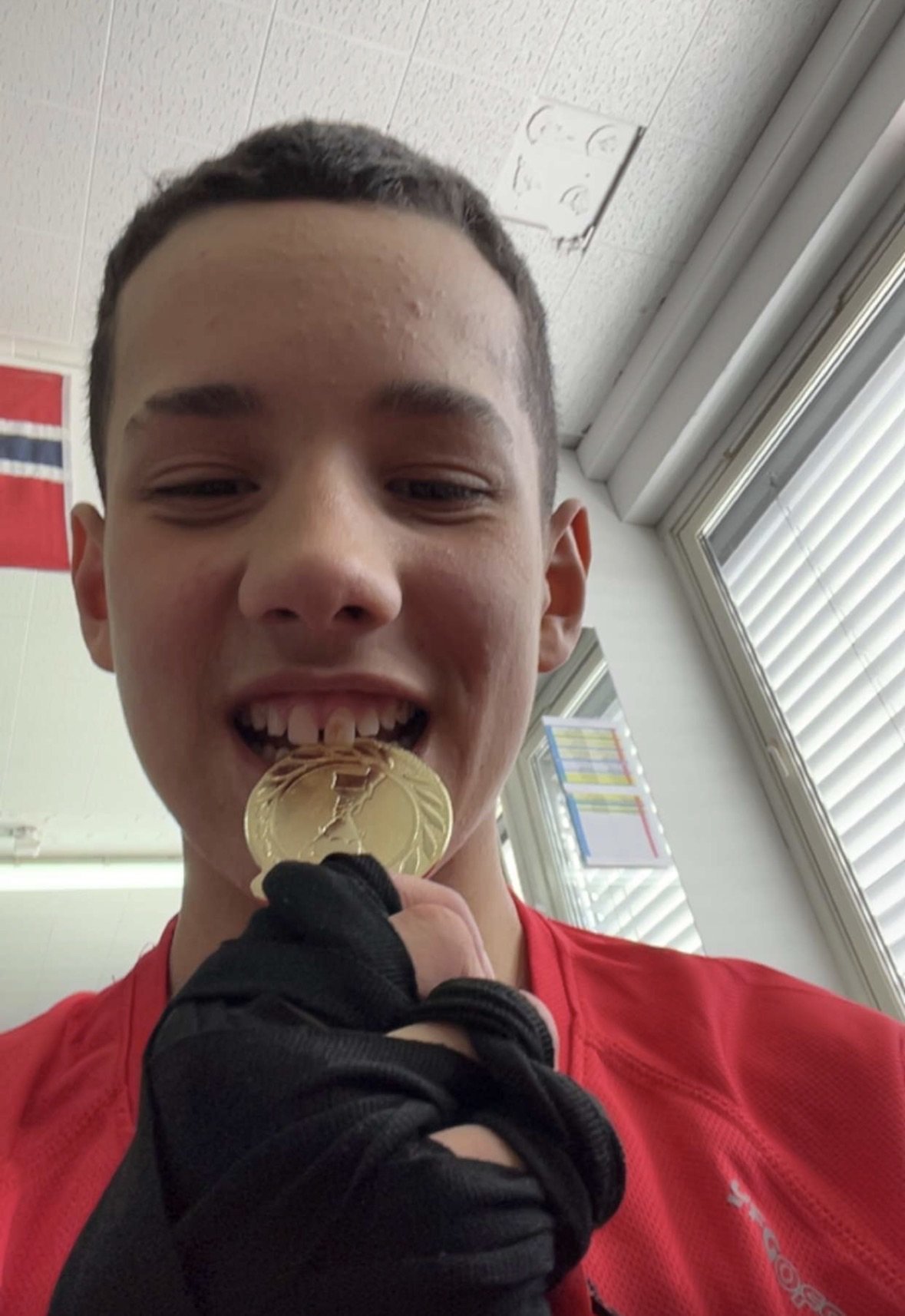Young person in red shirt proudly holding a gold medal in their mouth indoors