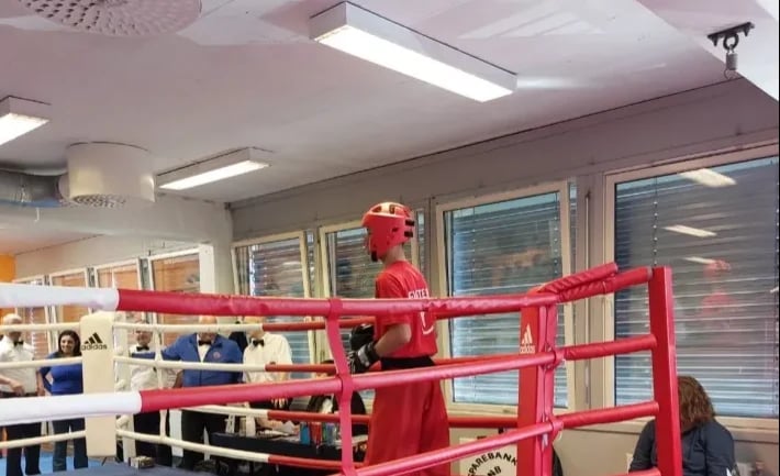 Red boxing ring with figure inside in classroom with desks, windows, and fluorescent lighting