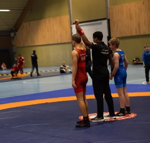 Wrestling referee raises the hand of a young competitor in red uniform after match victory at an indoor gymnastics facility