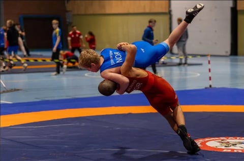 Young wrestler in blue uniform performing a throwing technique on opponent in red at indoor wrestling competition