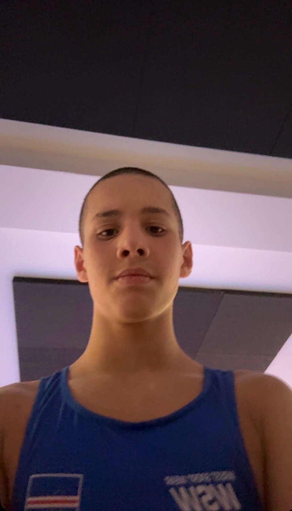Portrait of a young person with short hair wearing a blue tank top, photographed indoors against a light-colored wall.